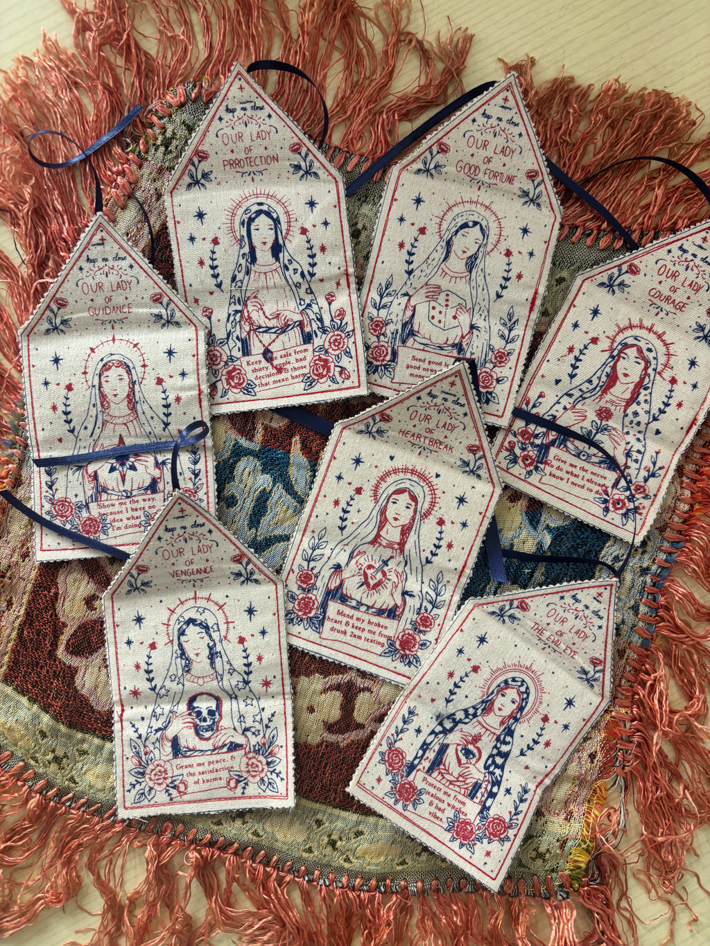 A set of seven small talismans featuring semi religious illustrations of the female deities, printed in red and blue on a cream background, arranged on a patterned rug with coral fringe.