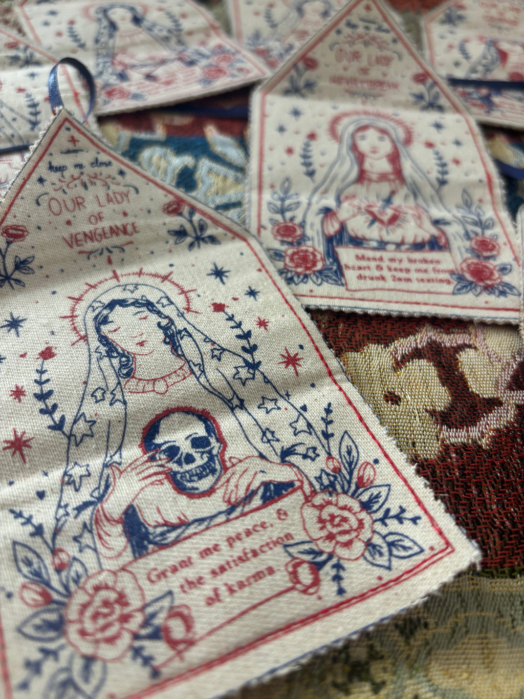 A close-up of several small fabric amulets featuring semi religious illustrations of Our Lady of Vengeance, with red and blue screen print on an off-white woven background.