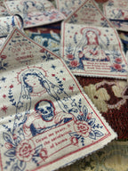 A close-up of several small fabric amulets featuring semi religious illustrations of Our Lady of Vengeance, with red and blue screen print on an off-white woven background.