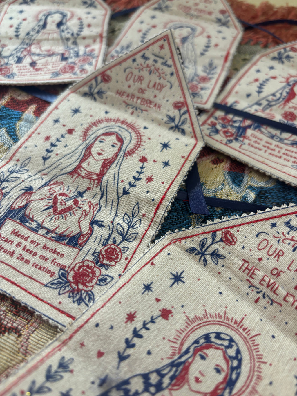 A close-up of several fabric prayer flags or bookmarks featuring religious illustrations and printed text, arranged on a patterned textile background.