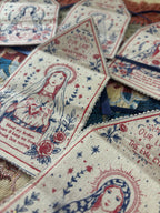A close-up of several fabric prayer flags or bookmarks featuring religious illustrations and printed text, arranged on a patterned textile background.