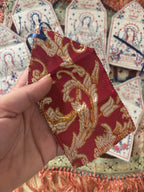 A hand holds a small red embroidered fabric tag with gold and white ornamental patterns and a blue ribbon loop, with other similar tags visible in the background.