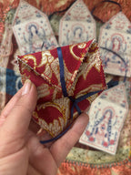 A hand holds a small folded red and gold embroidered fabric tag with a blue ribbon, with several similar hanging fabric tags displayed in the background.