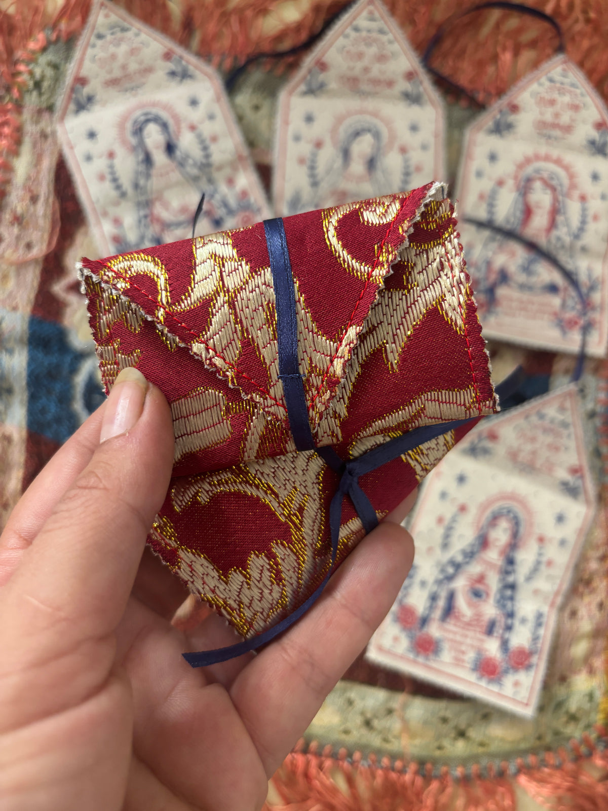 A hand holds a small folded red and gold embroidered fabric tag with a blue ribbon, with several similar hanging fabric tags displayed in the background.