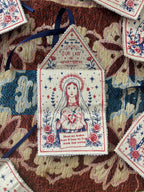 A close-up of a decorative hand printed fabric amulet, featuring a religious illustration of a female saint with a broken heart and the text 'Our Lady of Heart Break.' The talisman is surrounded by other patterned fabric pieces and dark blue ribbons on a textured multicolored textile background.