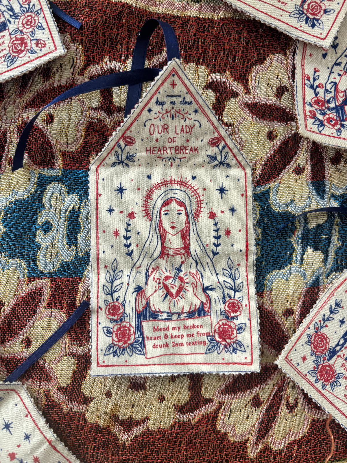A close-up of a decorative hand printed fabric amulet, featuring a religious illustration of a female saint with a broken heart and the text 'Our Lady of Heart Break.' The talisman is surrounded by other patterned fabric pieces and dark blue ribbons on a textured multicolored textile background.
