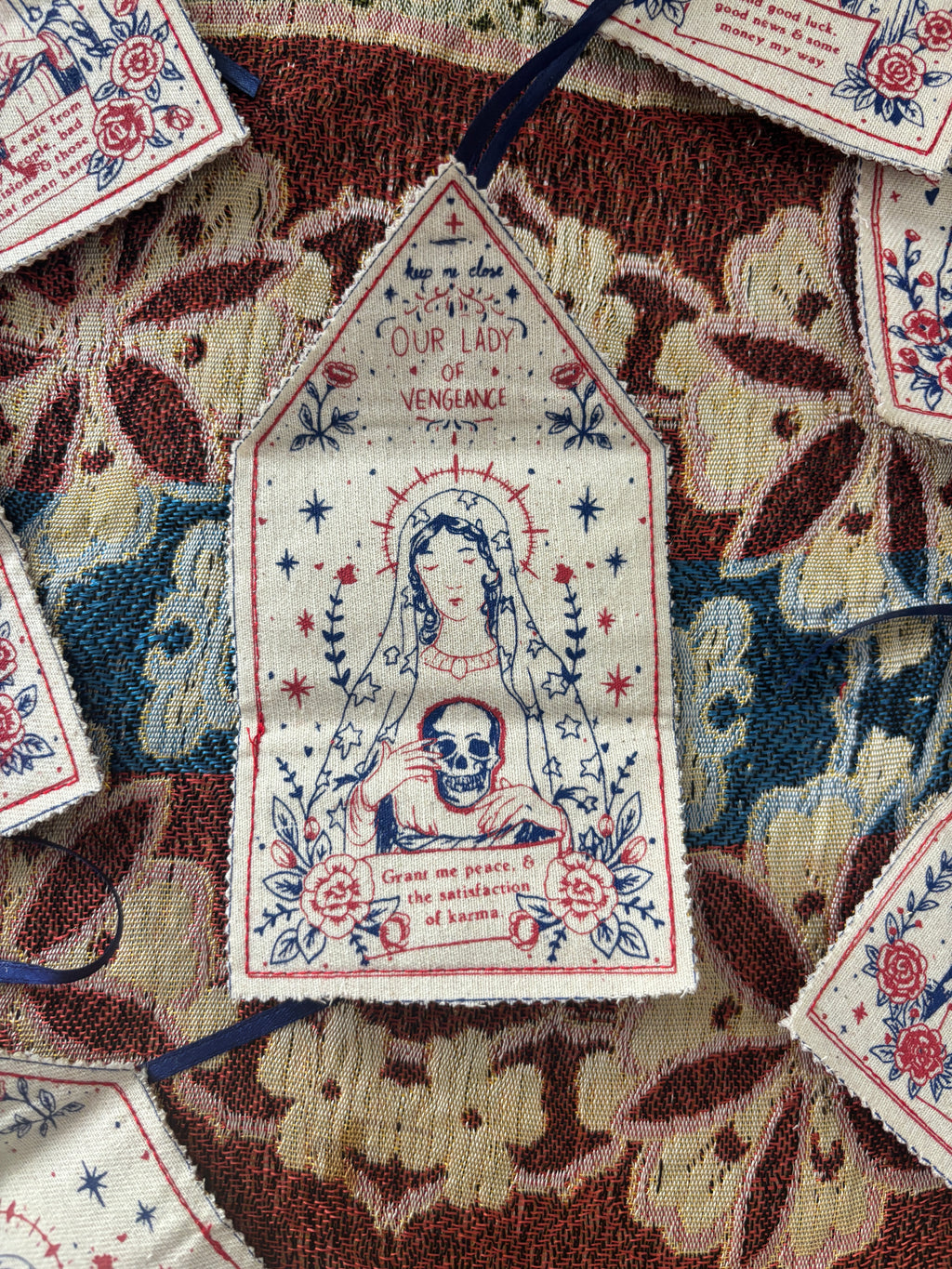 A close-up of a decorative embroidered fabric amulet laid on a patterned textile background. The amulet has a hand printed illustration of a robed figure holding a skull, surrounded by floral and star motifs, with the text 'Our Lady of Vengeance' and additional prayer text.
