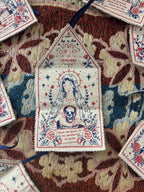 A close-up of a decorative embroidered fabric amulet laid on a patterned textile background. The amulet has a hand printed illustration of a robed figure holding a skull, surrounded by floral and star motifs, with the text 'Our Lady of Vengeance' and additional prayer text.