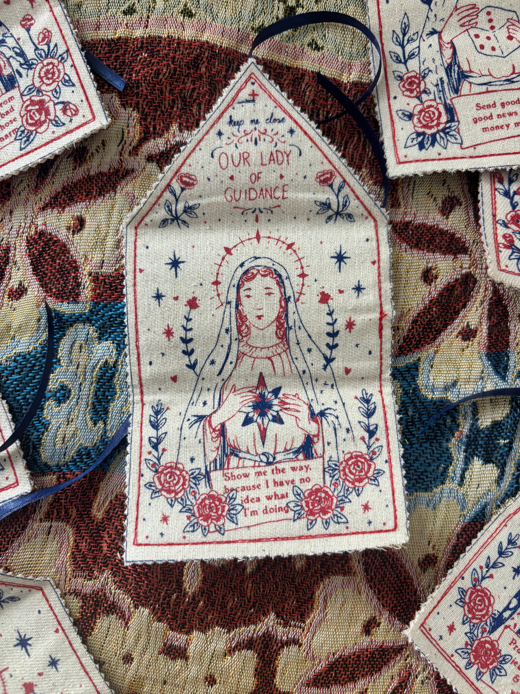 A close-up of a handmade talisman, featuring a religious illustration of Our Lady of Guidance with red and blue print on a cream fabric background. The patch is displayed on top of a patterned textile.