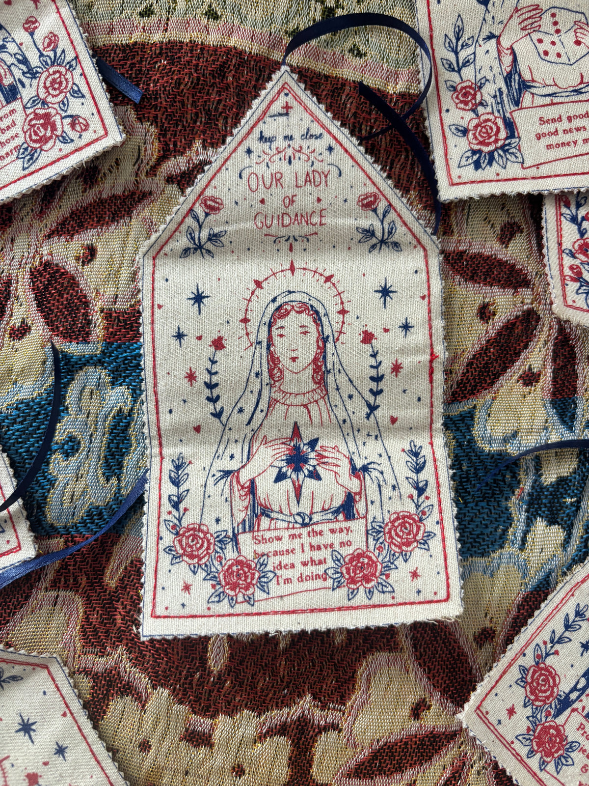 A close-up of a handmade talisman, featuring a religious illustration of Our Lady of Guidance with red and blue print on a cream fabric background. The patch is displayed on top of a patterned textile.