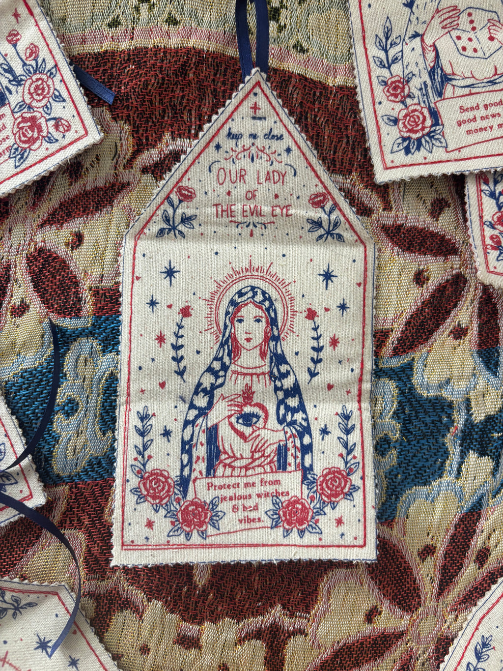 A close-up of a small embroidered fabric ornament shaped like a house, featuring Our Lady of the Evil Eye with red, blue, and cream print, displayed on a patterned textile background.