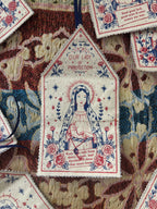 A close-up of embroidered fabric tags laid on a patterned textile background. The central tag shows a religious design with a robed figure, floral embroidery, stars, and the words 'Our Lady PROTECTION' 