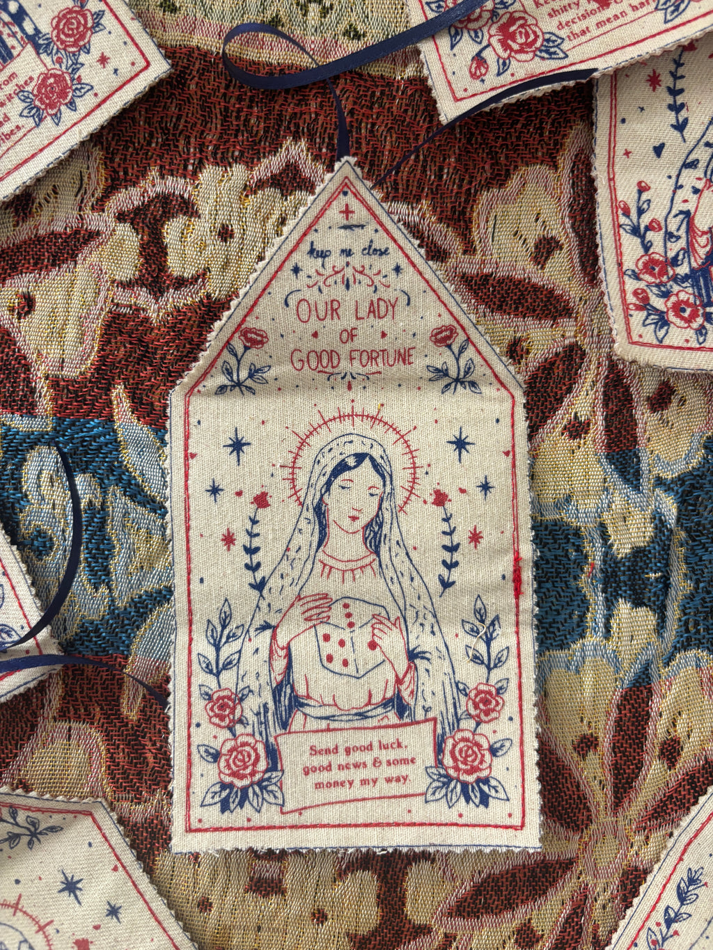 A close-up of a decorative embroidered fabric patch featuring Our Lady of Good Fortune, surrounded by other matching embroidered patches on a patterned textile background.