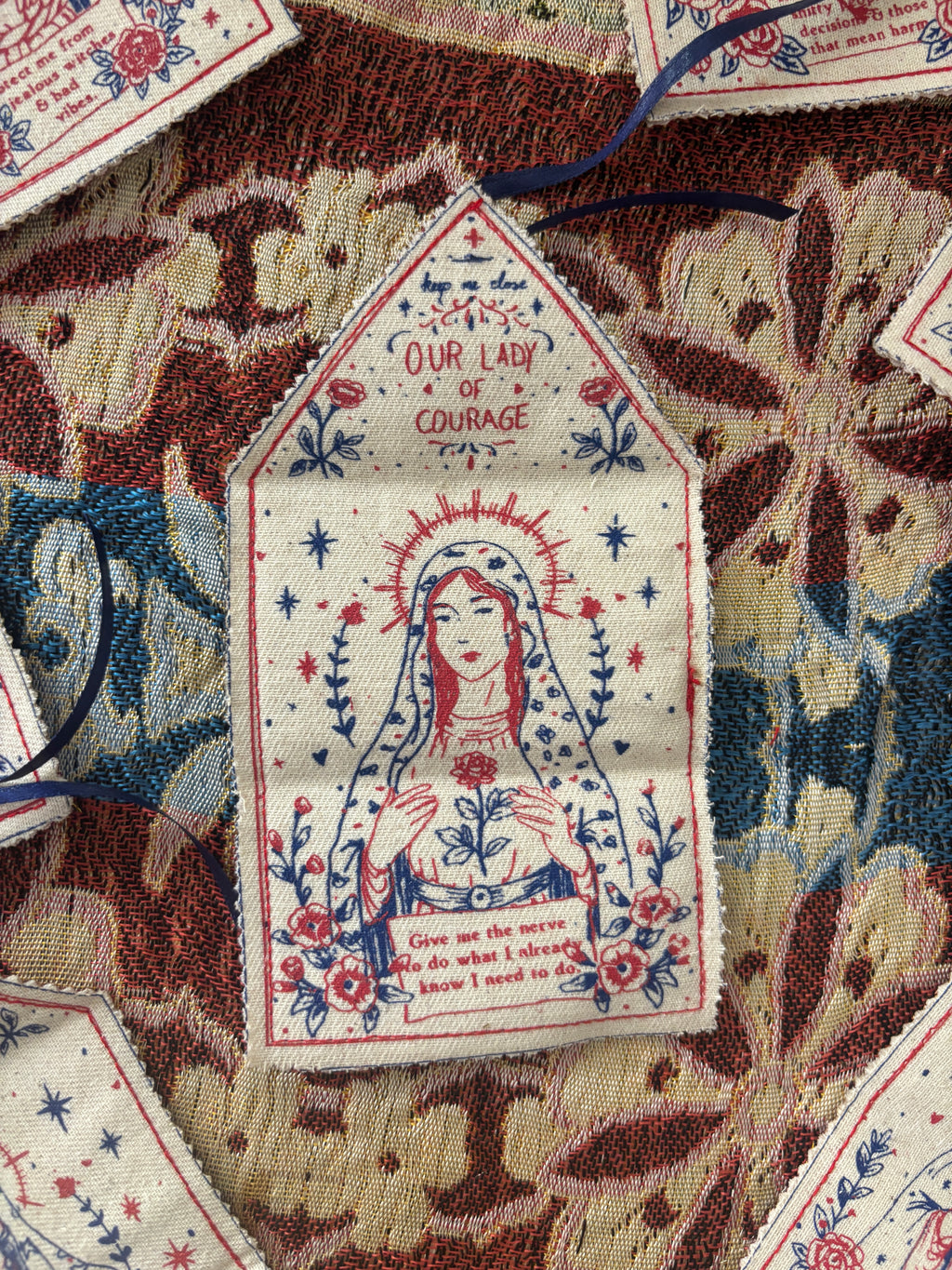 A close-up of a small embroidered fabric tag shaped like a house, featuring Our Lady of Courage in red, blue, and cream embroidery, surrounded by floral patterns and placed on a multicolored textile background.