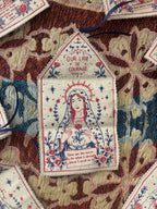 A close-up of a small embroidered fabric tag shaped like a house, featuring Our Lady of Courage in red, blue, and cream embroidery, surrounded by floral patterns and placed on a multicolored textile background.
